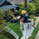 Electrician in a bucket lift repairing power lines from a utility pole in a suburban neighborhood.
